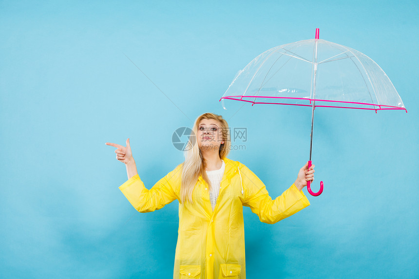 金发女人穿着黄色雨衣,着透明的雨伞,检查天气否下雨,用手指指着电脑图片