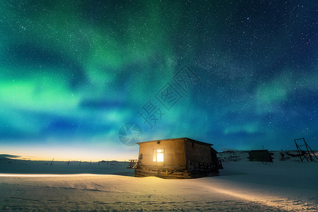 特里伯克北极光覆盖着古老的小房子,窗户发出光俄罗斯泰伯卡的北极光蓝天上星星绿色的北极光夜间冬季景观与极光,建筑背景