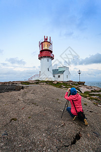 女游客历史上红色白色灯塔拍照,岩石海岸边缘,南挪威,林德斯尼斯FYR信标挪威林德内斯灯塔的游客背景图片