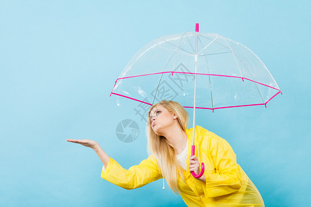 金发女人戴着黄色雨衣,着透明的雨伞检查天气,如果下雨的话背景图片