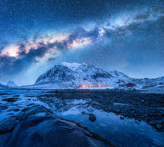 雪上的银河覆盖了冬天的山脉岩石海滩挪威洛福腾群岛着蓝色星空水石头雪岩明亮的星系城市灯光的景观背景图片