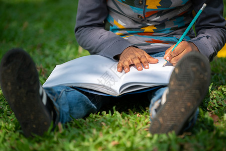 公园里的小孩坐在绿草地上时用铅笔在笔记本上写作图片