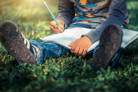 小孩坐在公园草地上用铅笔在笔记本上写作图片