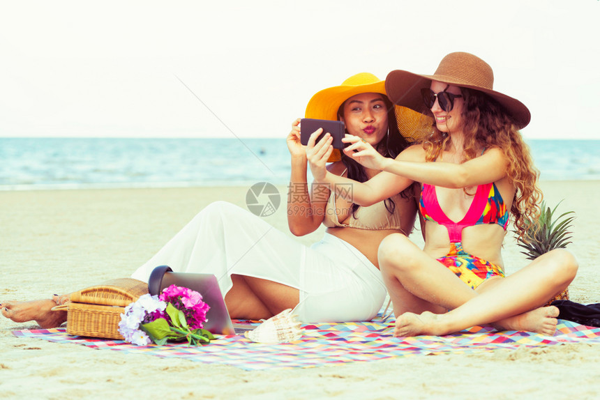 夏季在热带沙滩上穿着比基尼的快乐女子一起用手机自拍图片