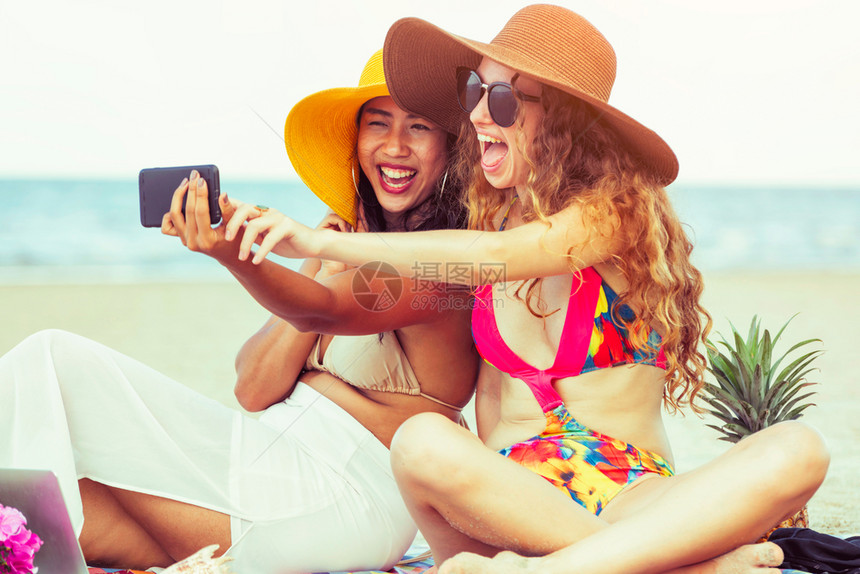 夏季在热带沙滩上穿着比基尼的女子一起用手机开心自拍图片