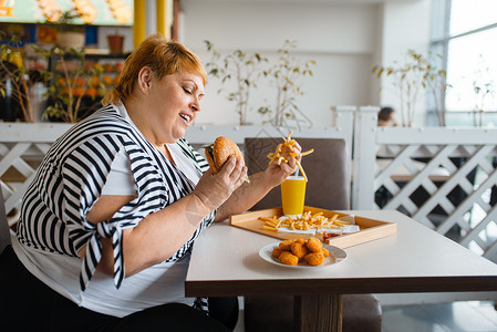 在快餐厅吃高卡路里食品的胖女人在餐桌上吃垃圾晚饭的超重女肥胖问题在餐厅吃高卡路里食品的胖女人背景