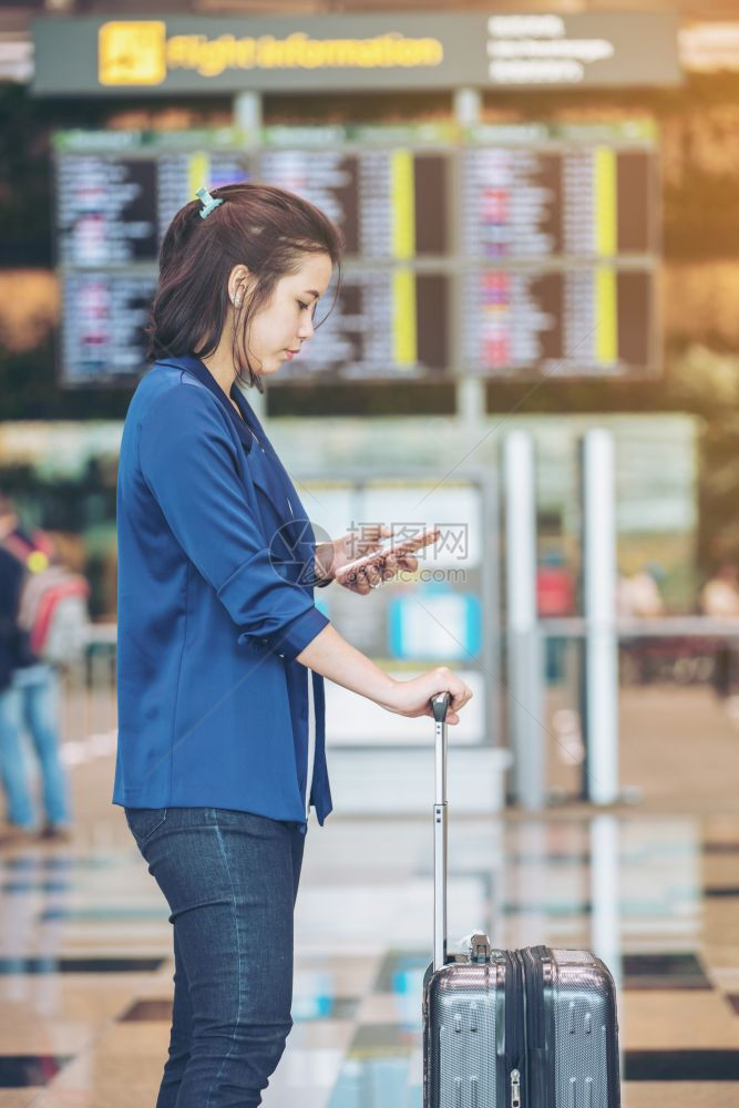 在国际机场使用智能电话等待报到时间的飞行信息委员会附近携带行李的年轻旅游女客图片