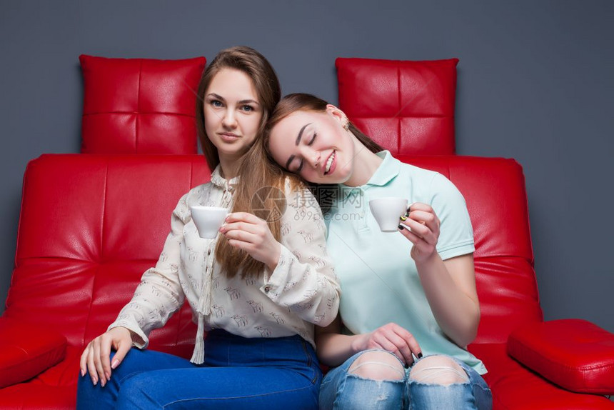 两个女孩端着咖啡一起坐在红色的皮沙发上图片