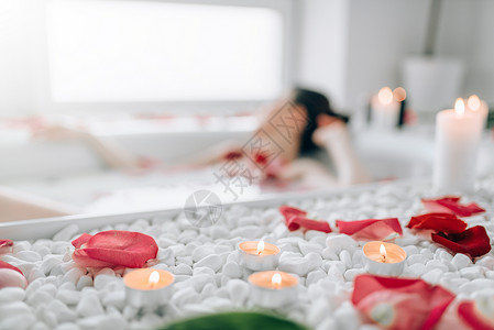 女人躺在浴池里身上沾着泡沫玫瑰花瓣烧蜡烛完全放松浴室里浪漫背景图片