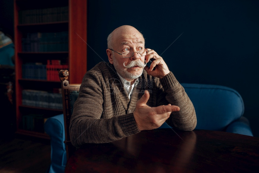 老年男子在家庭办公室用移动电话说在起居室留胡子的成年老人商在家庭办公室用移动电话说的老年男子图片