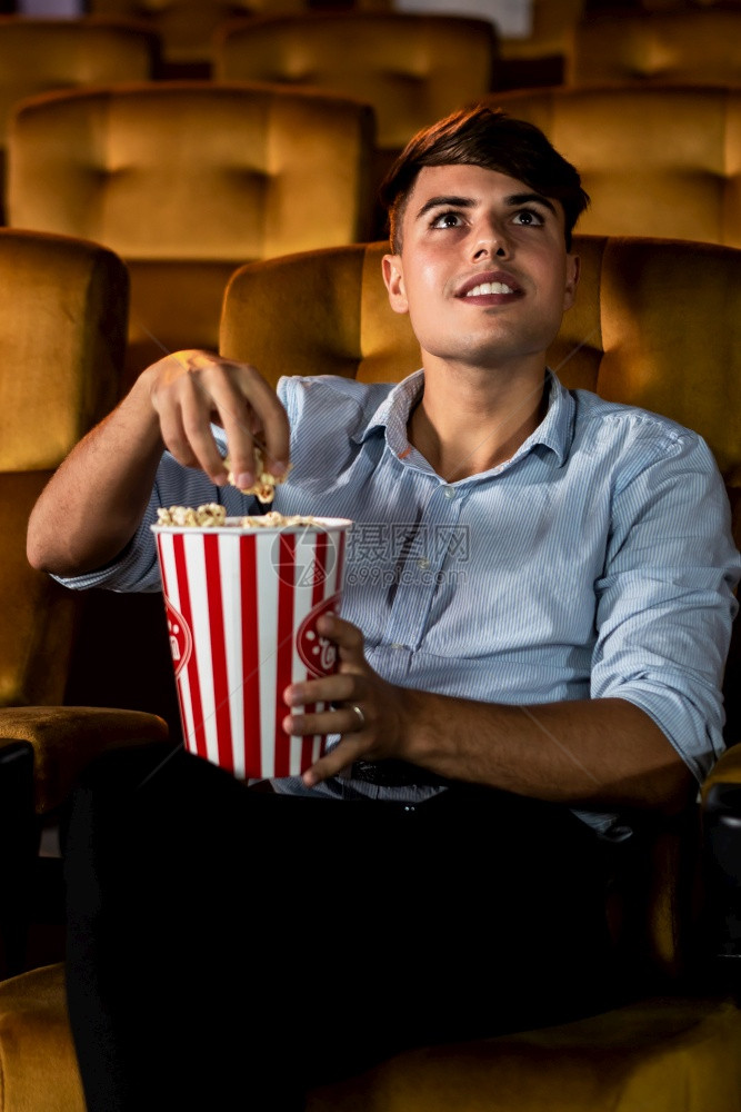 一个年轻男人在电影院看时微笑着享受爆米花和图片