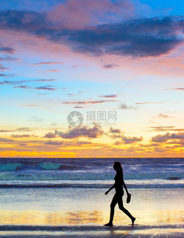 日落时在海滩上走的女子赤脚手拿着鞋子图片