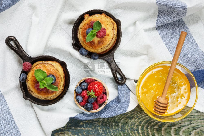 煎饼在铸铁煎锅里新鲜的浆果和蜂蜜放在一个玻璃碗里背景是用亚麻桌布覆盖的旧木板图片