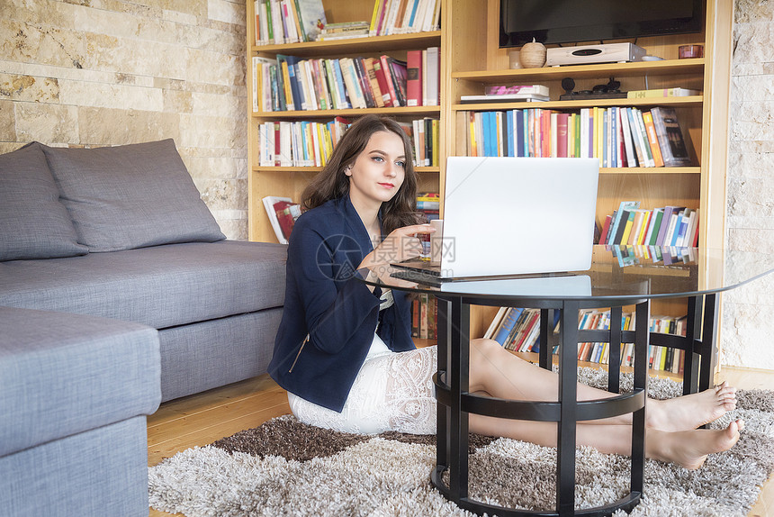 穿着优雅的年轻美女坐在她家舒适的地毯上享受咖啡同时使用笔记本电脑图片