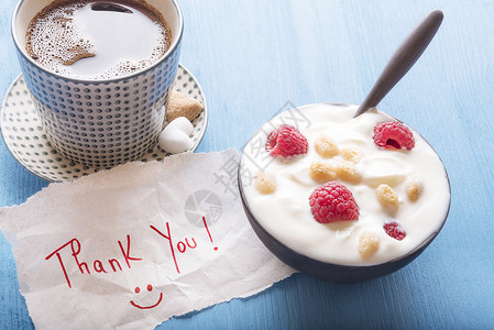 笑脸酸奶加上酸奶草莓和麦片一杯热咖啡和张感谢信面带微笑的孔在清晨阳光下放在一张蓝色的木桌上背景