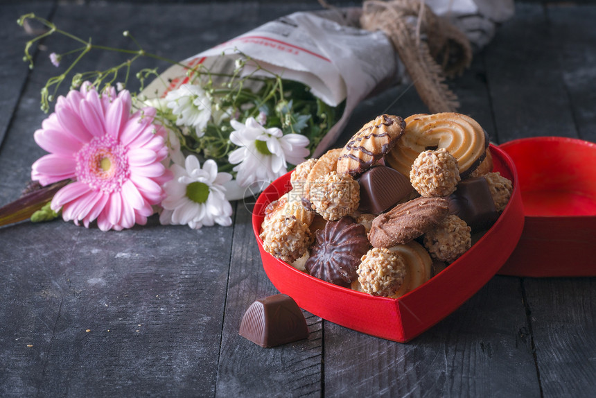 红盒装满饼干巧克力和花束包在报纸背景旧木制桌子上图片