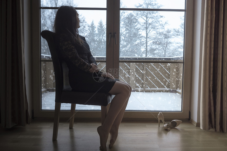 穿着黑裙子的优雅年轻黑发女人坐在椅子上放松看着大玻璃门的雪花图片