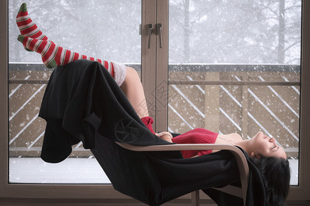 穿着短红色裙子和长袜的年轻感女人坐在椅子上倒着放松站在玻璃门前外面在下雪背景图片