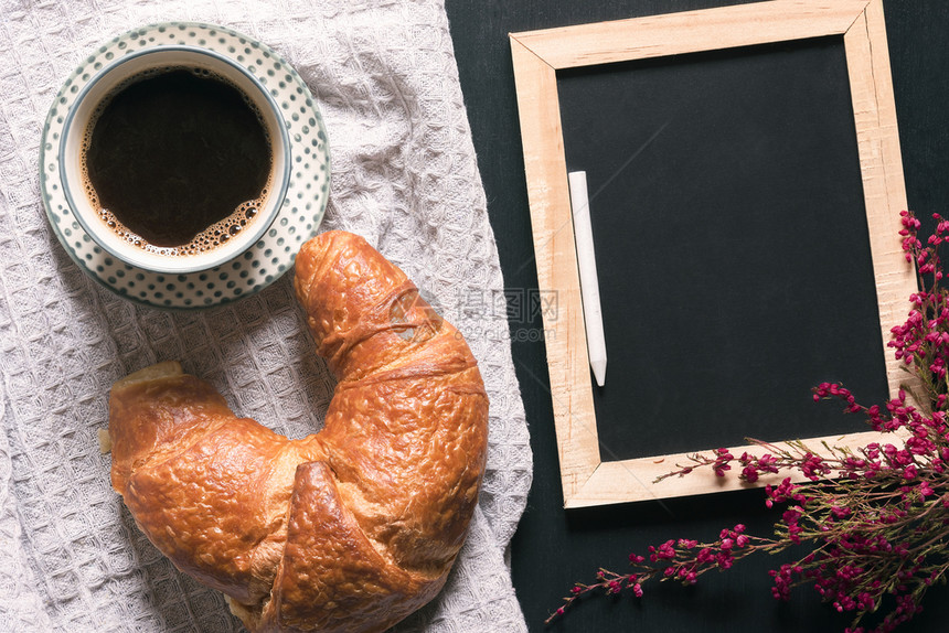 早餐桌有一杯咖啡和角面包放在旧式厨房毛巾上在黑桌子一个空的板上面有鲜花环绕的空粉笔板图片