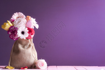 挂着花朵的玫瑰郁金香和雪贝拉的生日卡包裹在陈年的帆布袋里放在粉红色的木桌和紫背景上背景图片