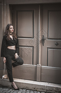 自信的年轻女人穿着优雅的现代黑衣靠在墙上站一扇大旧木门前抬头看图片