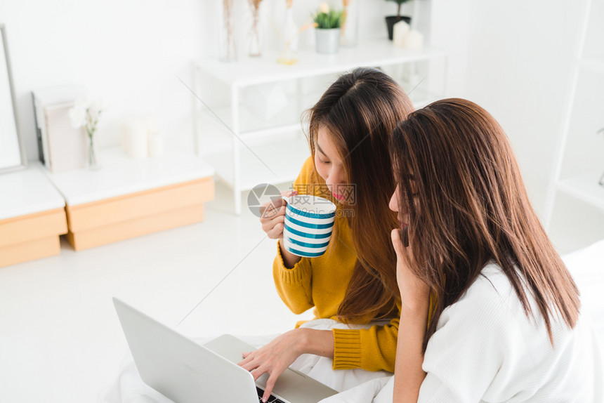 lgbt女快乐夫妇坐在床上拥抱使用笔记本电脑同时在家中一起喝热咖啡杯lgbt女夫妇一起在室内概念图片