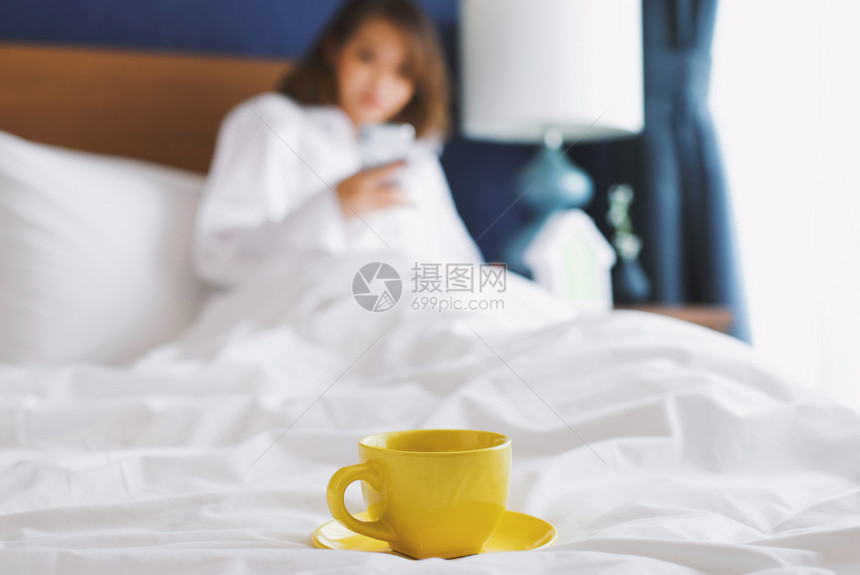 早上在床用智能手机与妇女一起关注黄色咖啡或茶杯图片