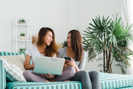 坐在沙发上用电脑和手机在家中客厅购买lgbt女夫妇一起在室内的概念图片