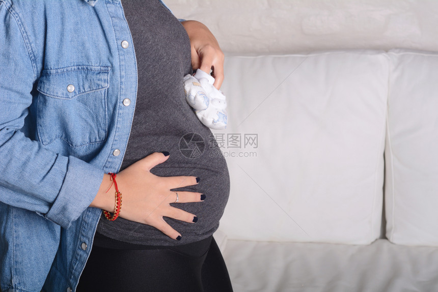 美丽的年轻孕妇将婴儿靴子放在肚子上图片
