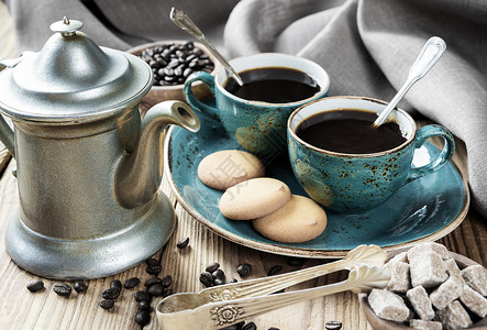 两杯黑色咖啡饼干和古董泡菜咖啡壶用旧木制桌布糖片和咖啡豆包着图片