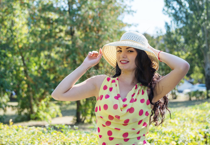 穿着轻丝裙的美丽长发棕黑美女带有波尔卡圆点和夏日自然背景下的草帽图片