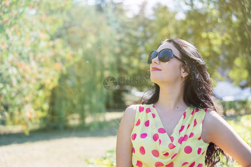 美丽的长头发棕黑穿着轻丝绸礼服带波尔卡圆点和太阳眼镜在夏日自然的背景下太阳在光欢欣图片