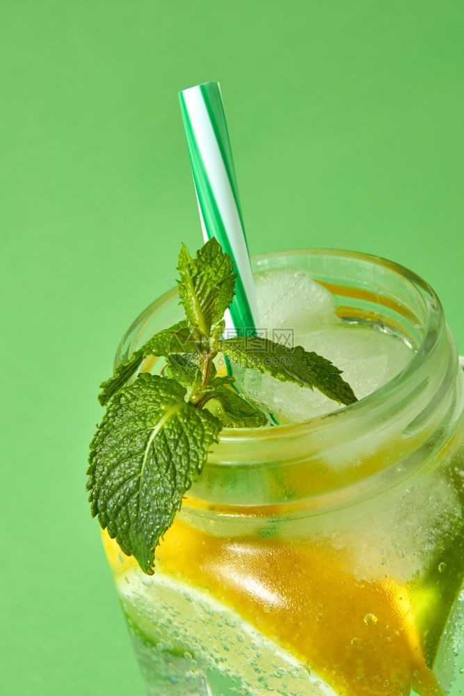一杯绿色薄荷放在玻璃罐中有柑橘片冰闪光水和彩色塑料吸管在绿色背景中隔离天然寒冷的夏季自制柠檬水一块绿色薄荷冷的自制柠檬在绿色上隔图片