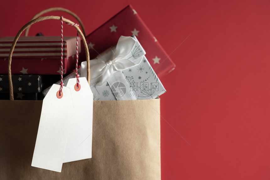 两个白色的无文字标签挂在纸袋上满是多彩的礼物盒红色背景上图片