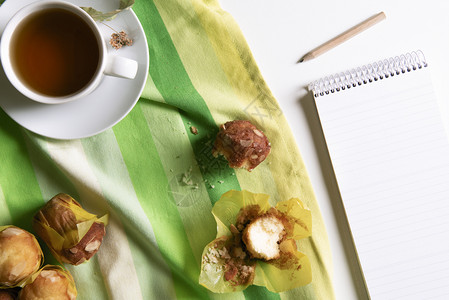 上面有一张早餐桌上面有一杯叶茶绿黄毛巾上有松饼白桌一本笔记图片