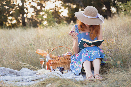 女孩坐在野餐篮子和面包葡萄酒眼镜和面包卷旁看书图片