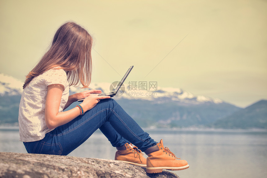 在山地和湖泊背景的岩石上坐着笔记本电脑的女孩徒步旅行者图片