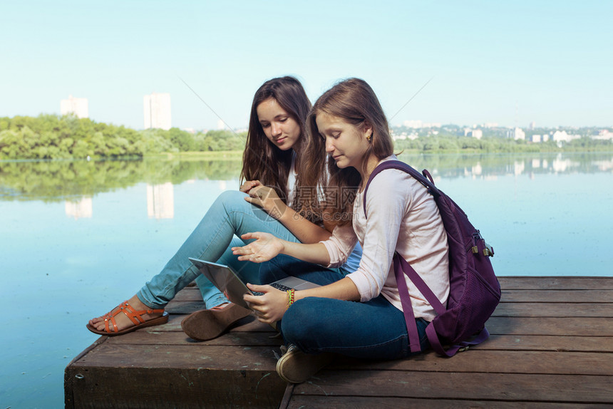 两个笑着的少女背包坐在河岸的码头上图片