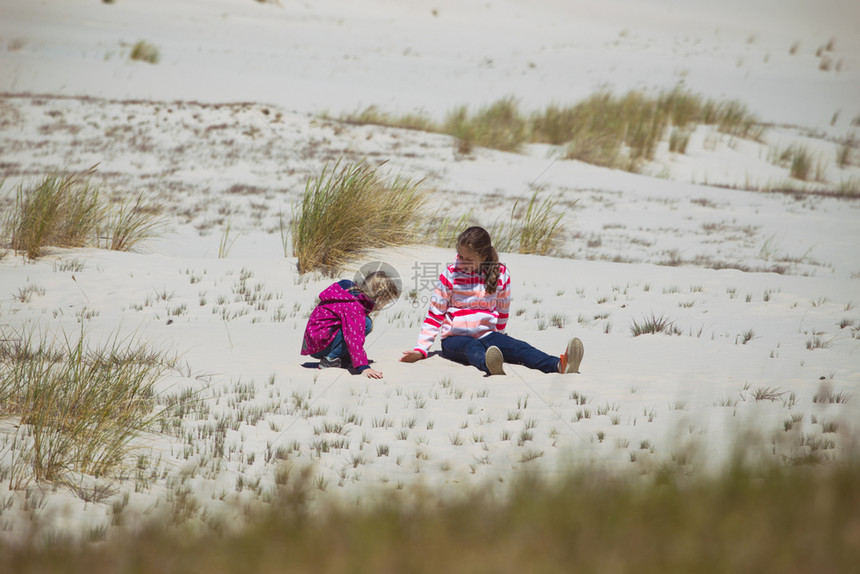 女孩姐妹们坐在沙滩上的沙丘和蓝天的背景下波罗的海图片