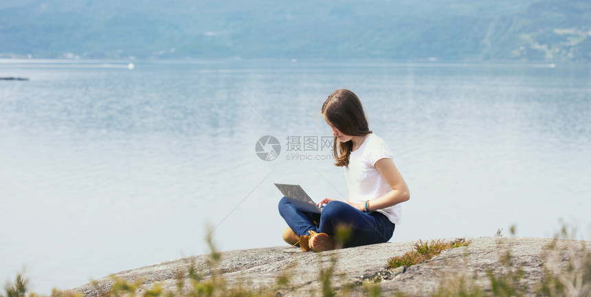 在山地和湖泊背景的岩石上坐着笔记本电脑的女孩徒步旅行者图片