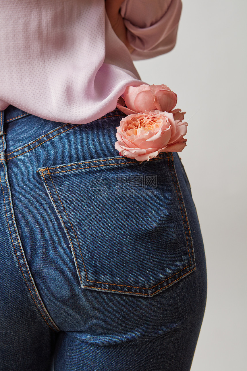 身穿牛仔裤的美丽女屁股鲜红的珊瑚玫瑰在浅灰色背景的后口袋里穿着牛仔裤和粉红色的衬衫图片