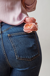 屁股口袋身穿牛仔裤的美丽女屁股鲜红的珊瑚玫瑰在浅灰色背景的后口袋里穿着牛仔裤和粉红色的衬衫背景