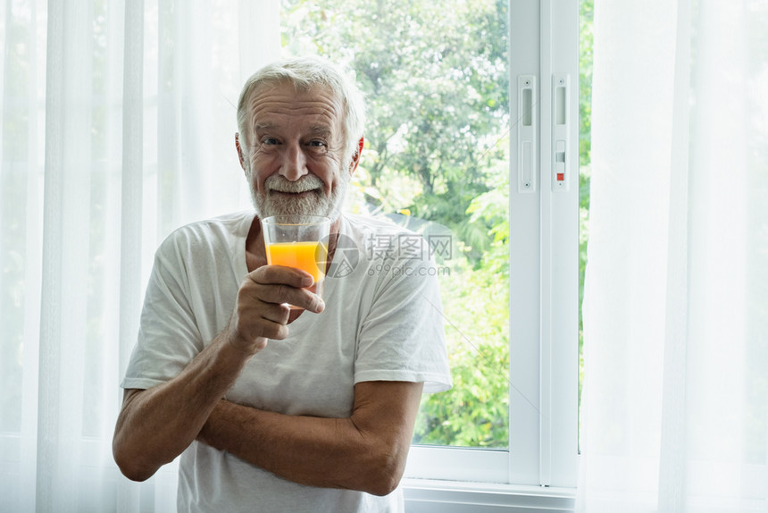 高级男子微笑并握着一杯橙子果汁摊在白色房间的窗户前图片