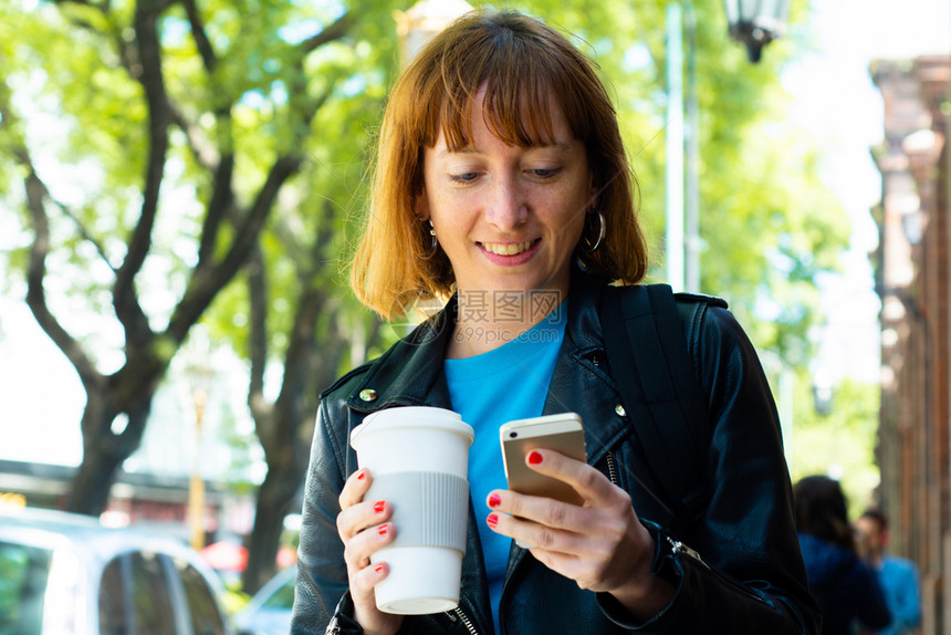 年轻红发女拿着咖啡和阅读信息在手机上户外图片