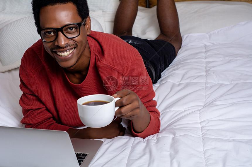 年轻美国男子看着相机用笔记本电脑在床上喝咖啡图片