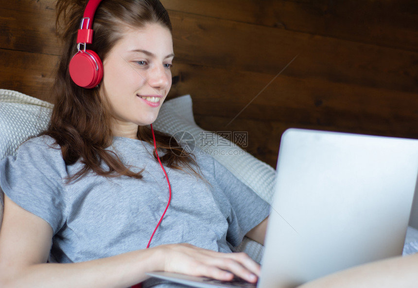 青年妇女在舒适的床上戴着耳机使用笔记本电脑图片