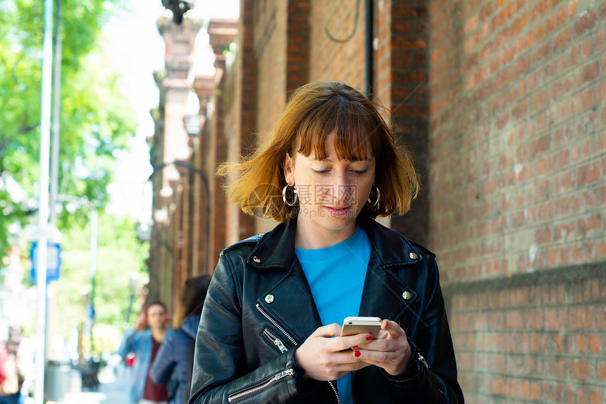 美丽的年轻红发女送信息智能手机在街上图片