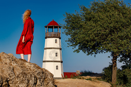 穿红裙子的女人站在灯塔附近的海滩上图片