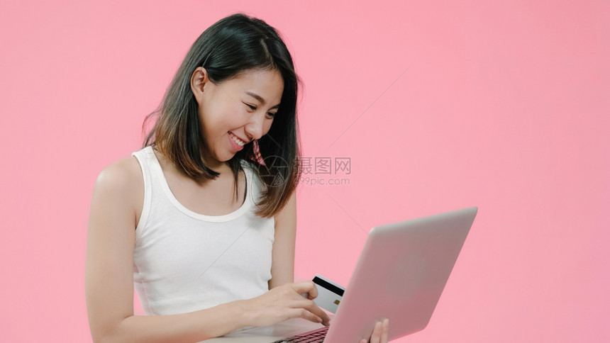 使用笔记本的年轻女用临时衣着信卡购买网上物透过粉红色背景工作室拍摄的电脑图片