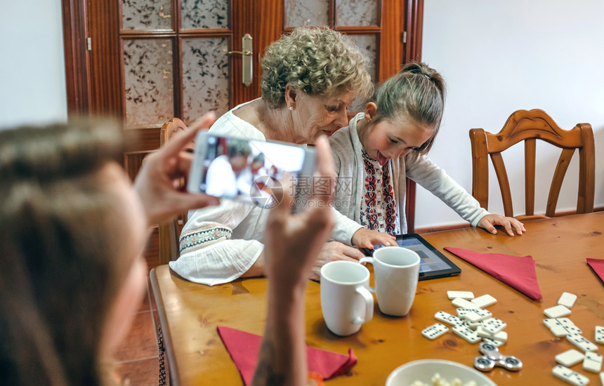 外祖母和孙女在平板上玩游戏而母亲则用智能手机拍照有选择地关注祖母和外孙女的背景图片